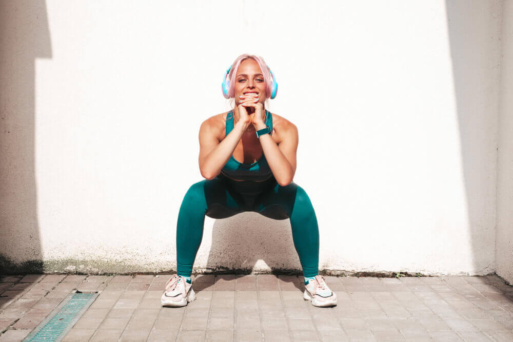 Fitness smiling woman in green sports clothing with pink hairShe doing squats Young beautiful model with perfect bodyFemale in the street near white wallCheerful and happy Outdoors in headphones
