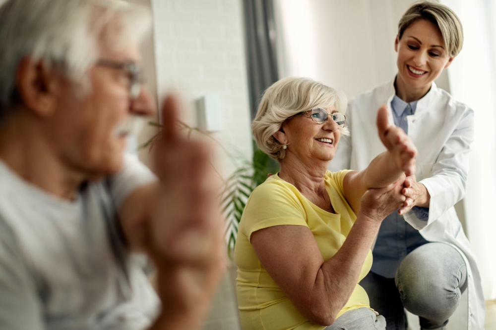 photo happy physiotherapist helping to senior couple with stretching exercises at home
