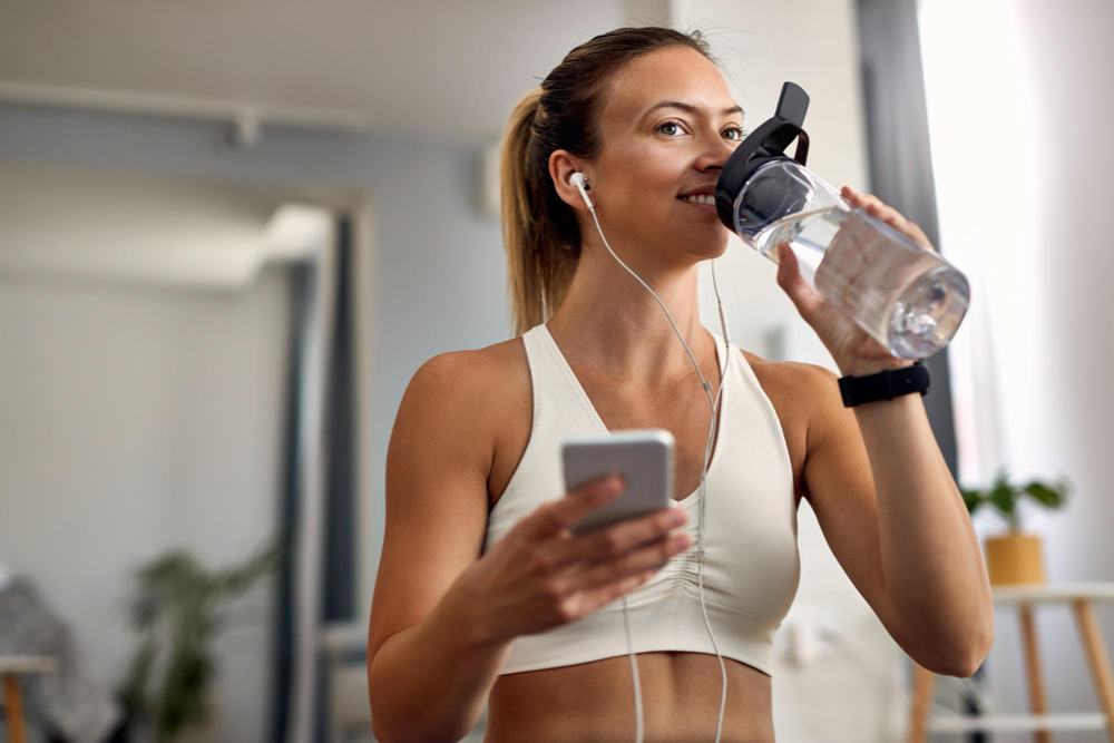 happy sportswoman using smart phone while having water break in the living room