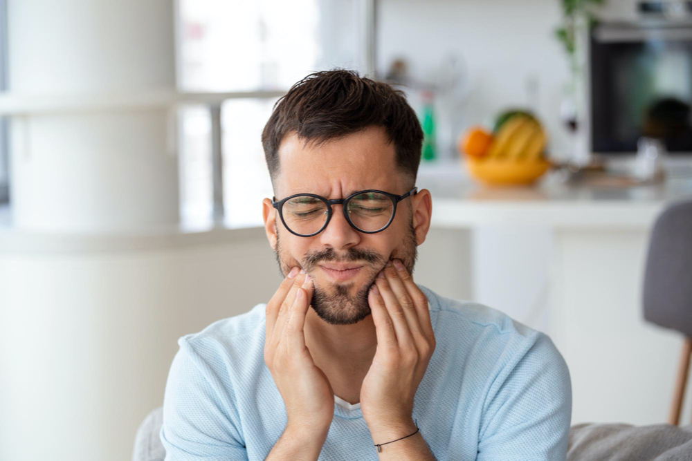 frustrated young man touching his cheek and keeping eyes closed while sitting on the couch at home