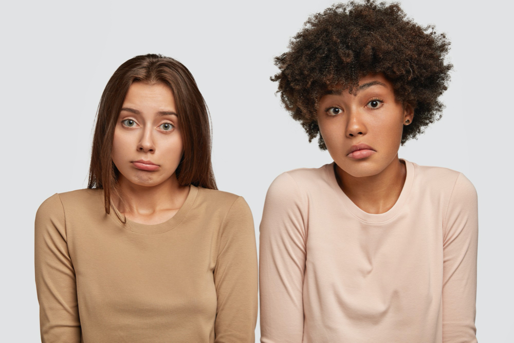 Puzzled women shrugg shoulders in bewilderment: beautiful European woman stand closely to African American female friend, cant make decision about something, isolated over white wall.