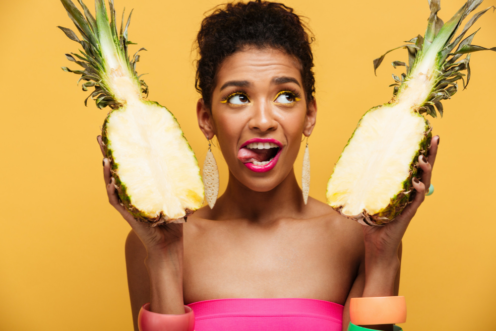 Amazing mulatto woman with colorful makeup looking upward and licking her lips while holding two parts of ripe appetizing pineapple isolated, over yellow
